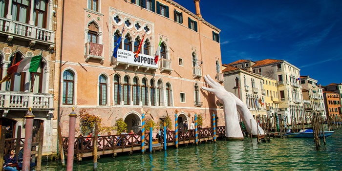 Large sculpture of two hands rising from a canal, supporting an old building in Venice, Italy. Bright blue sky and row of buildings in the background.