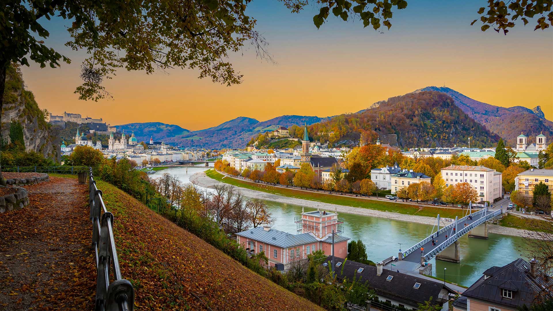 Beliebtes Reiseziel von Pilart-Air: Eine malerische Aussicht auf die Stadt Salzburg an einem Fluss, umgeben von Herbstbäumen und Hügeln, bei Sonnenuntergang. Eine Brücke überspannt den Fluss und auf der anderen Seite des Wassers sind verschiedene Gebäude zu sehen.