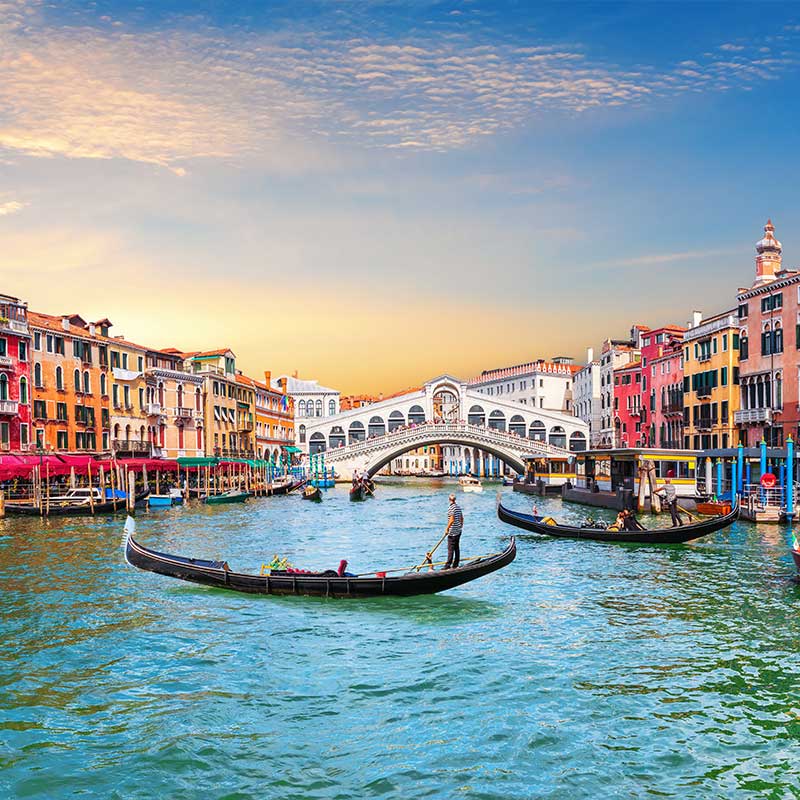 Gondeln auf dem Canale Grande in der Nähe der Rialtobrücke in Venedig, Italien, mit farbenfrohen Gebäuden und einem blauen Himmel im Hintergrund.