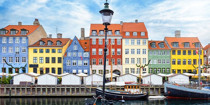 Colorful buildings line a waterfront with boats and a streetlamp in the foreground. The sky is partly cloudy.