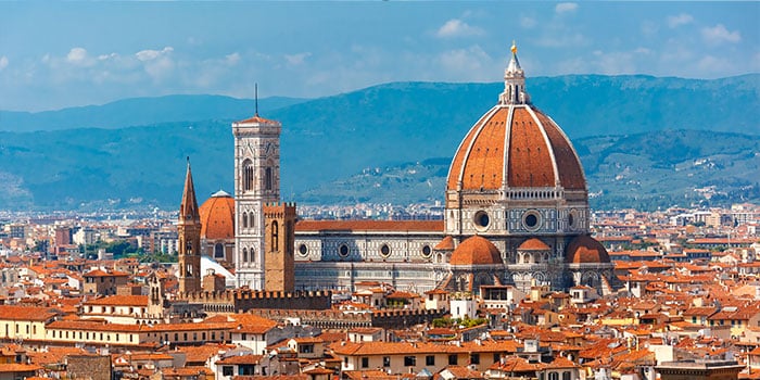 Panoramic view of Florence, featuring the Cathedral of Santa Maria del Fiore with its iconic red dome and bell tower, surrounded by city rooftops and distant hills.