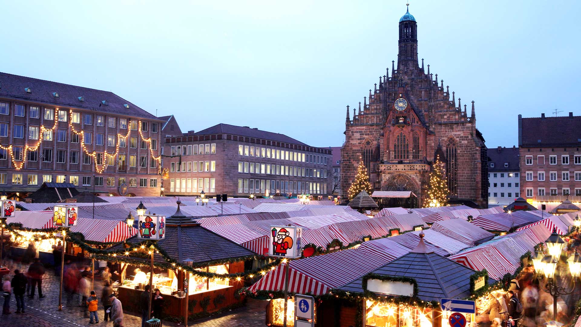 Christmas market stalls with red and white striped roofs are set up in front of a large, illuminated cathedral in the evening.