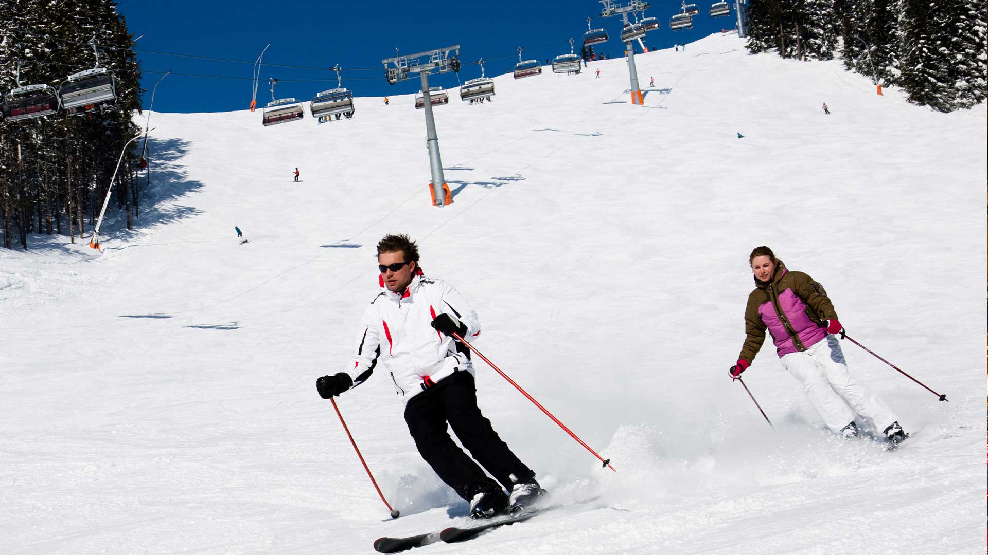 Zwei Personen fahren einen schneebedeckten Hang hinunter. Im Hintergrund sind Skilifte und weitere Skifahrer unter einem klaren blauen Himmel zu sehen.