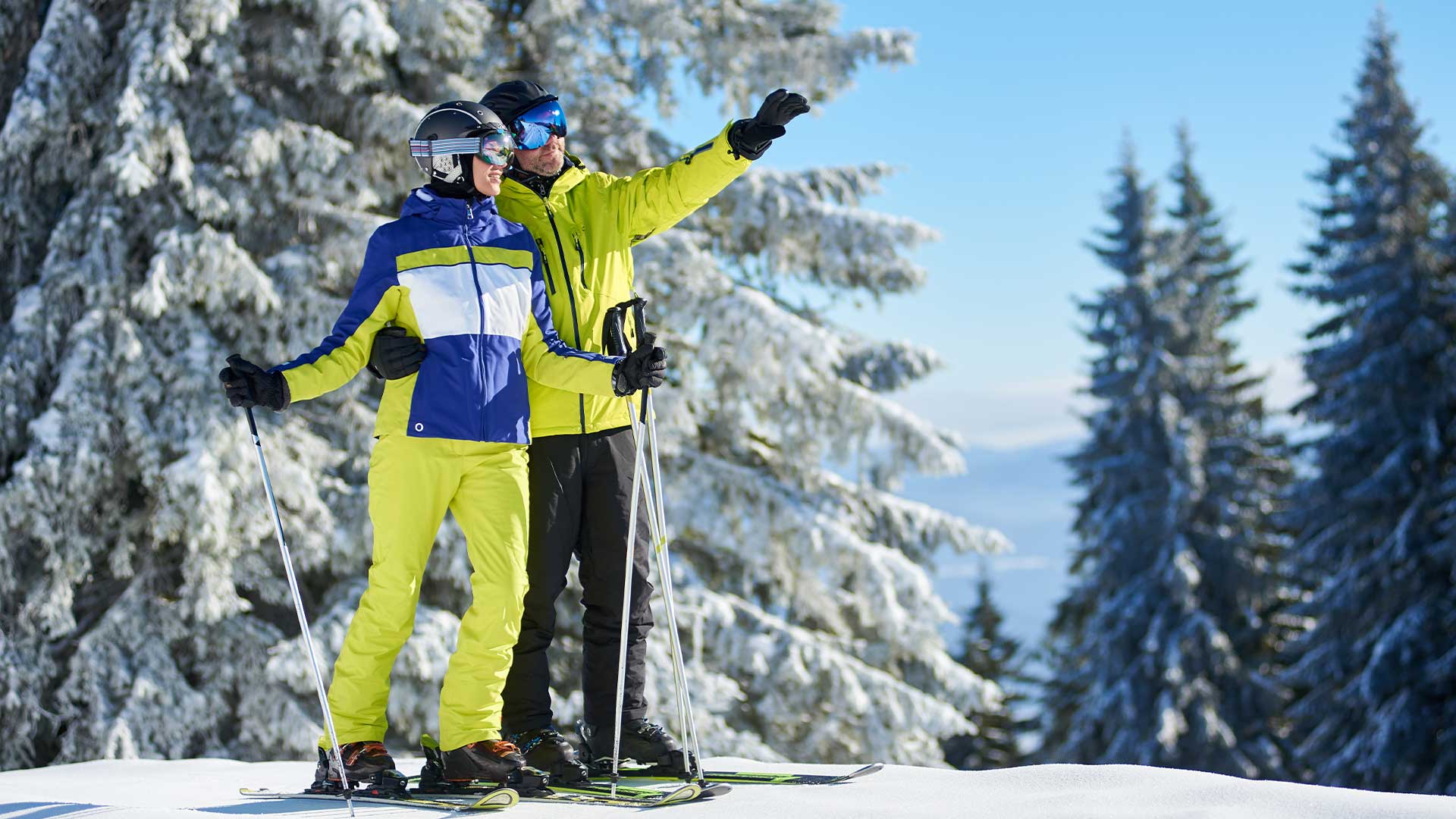 Zwei Skifahrer in bunter Ausrüstung stehen auf einem schneebedeckten Hang, einer zeigt in die Ferne. Im Hintergrund sind schneebedeckte Bäume zu sehen.