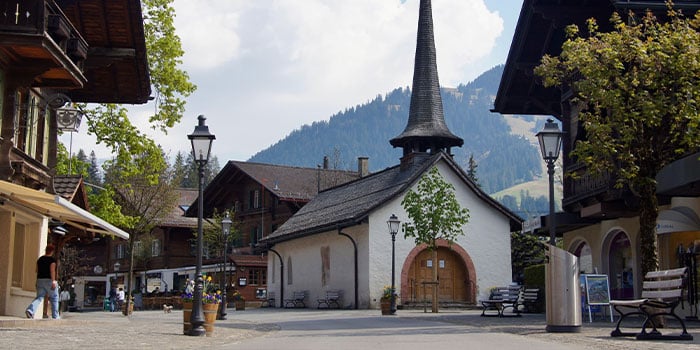 A quaint village street features a white chapel with a steep spire, surrounded by traditional wooden buildings, lampposts, and trees, with mountains in the background.