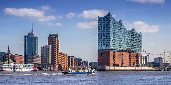 The image shows boats on the water in front of modern buildings, including the Elbphilharmonie with its wave-like glass structure, under a partly cloudy sky.