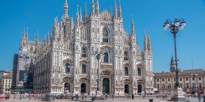 The image shows the Milan Cathedral, a Gothic-style church with intricate spires and a large facade, under a clear blue sky.