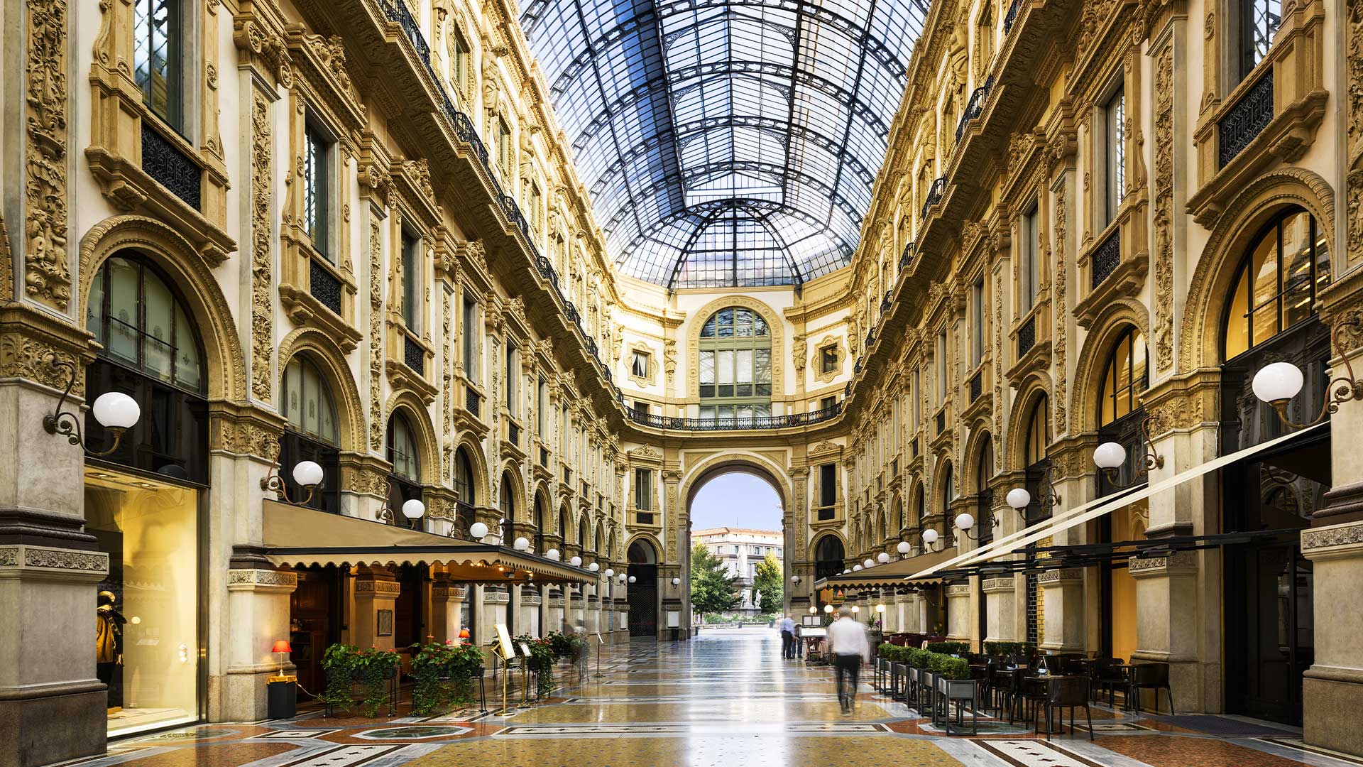 Elegant indoor shopping arcade with glass ceiling, ornate architecture, and decorative flooring. Tables and chairs line the sides, with a few people walking through the space.