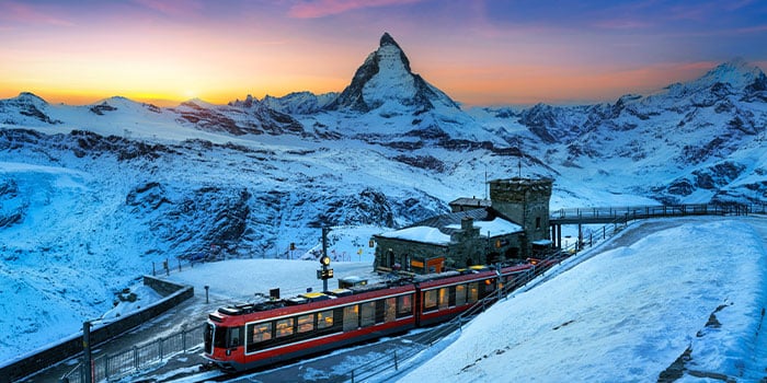 A red train is parked at a snowy mountain station during sunset, with the Matterhorn in the background under a clear sky.