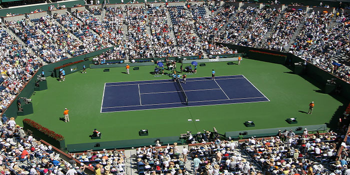 An aerial view of a tennis match in a stadium filled with spectators under a clear sky.