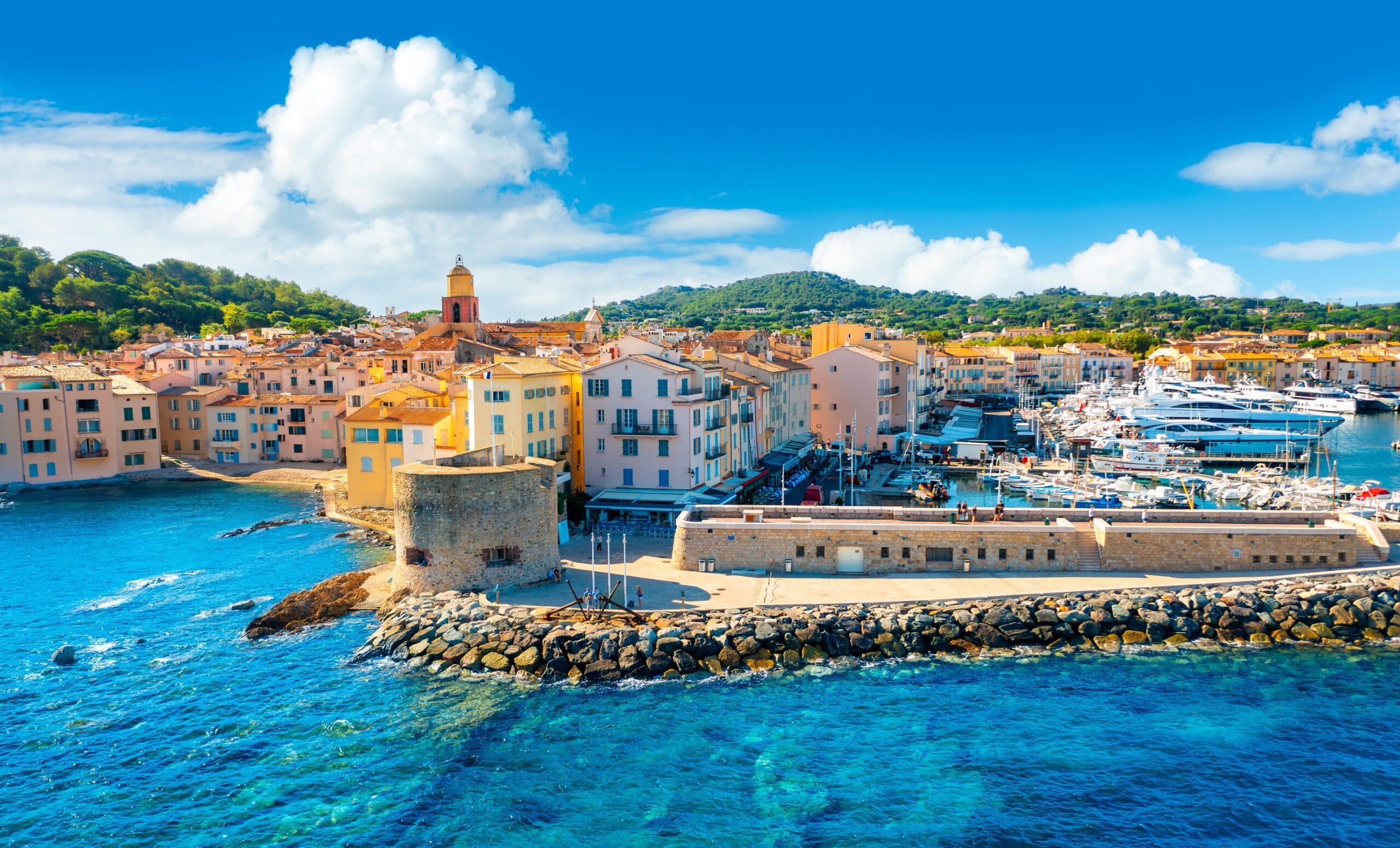 Eine malerische Küstenansicht von Saint-Tropez mit farbenfrohen Gebäuden, einem Glockenturm und festgemachten Booten in einem Yachthafen. Felsige Küste und klares blaues Meer unter einem sonnigen Himmel mit Wolken.